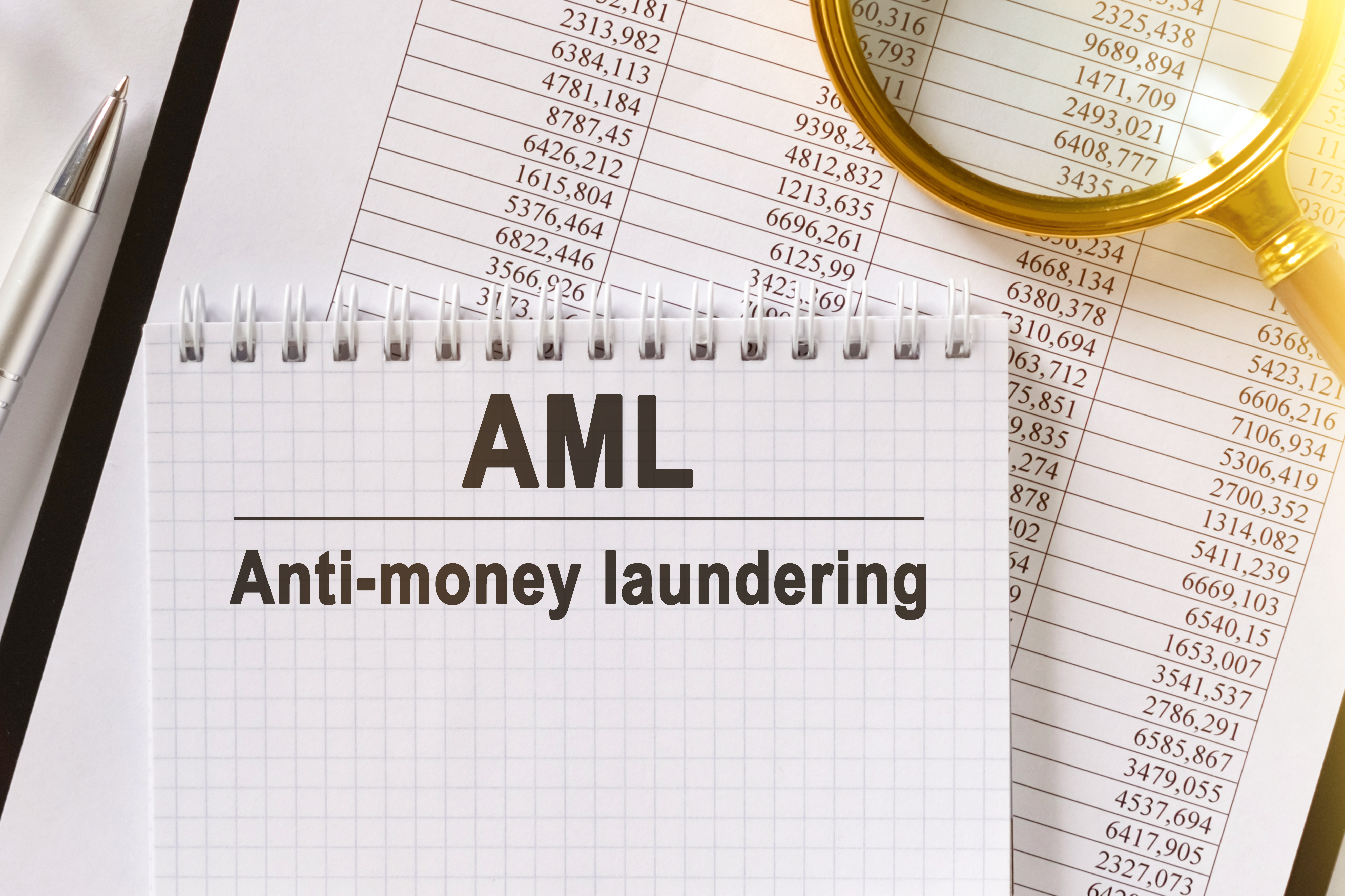 A sign that says 'AML - anti-money laundering' on it with a sheet of figures and a magnifying glass in the background.